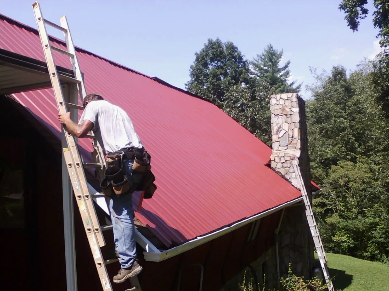 Roofer on a ladder beside a red metal roof and stone chimney — Soffit & Fascia Repair by Roofing Brothers in Elba